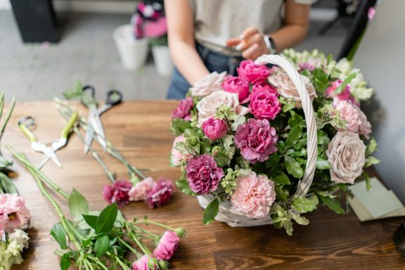 A person arranging a bouquet of flowers in a white basket on a wooden table.