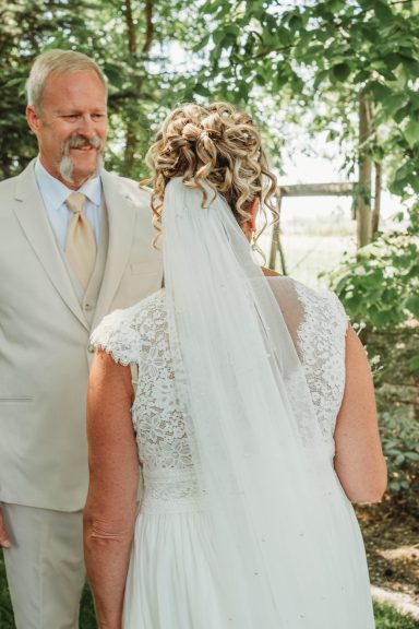 First Glance Magic A bride in a white dress and veil stands facing a groom in a light suit, surrounded by greenery.