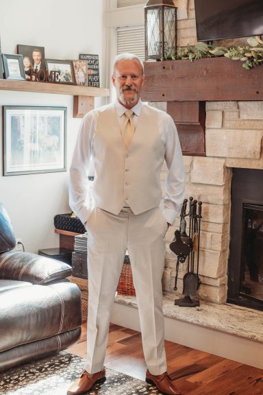 Ready for “I Do” Man in a light-colored suit stands confidently near a fireplace in a cozy living room.