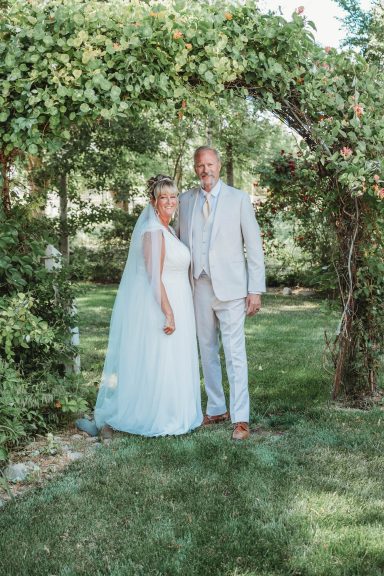 Under the Arbor Couple in wedding attire standing under a floral arch in a garden.