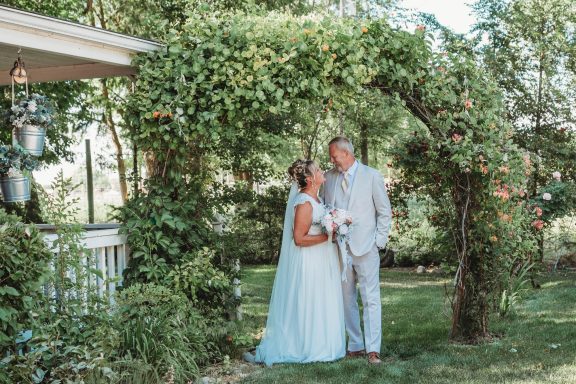 Love Under the Arbor Bride and groom stand happily under a floral archway in a sunny outdoor setting.