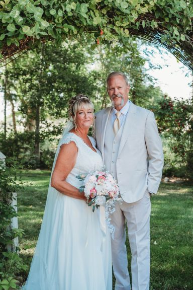 Framed in Love Bride in a light blue gown and groom in a light gray suit, standing under an archway.