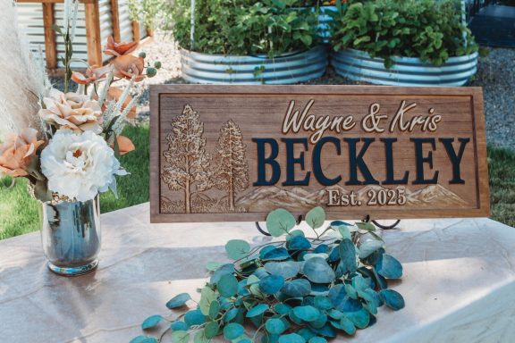 Custom Details, Picture-Perfect Wooden sign with "Wayne & Edna Beckley" engraved, surrounded by flowers and greenery.