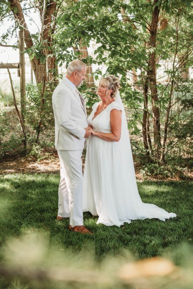 Among the Trees Bride and groom holding hands, surrounded by greenery, in a serene outdoor setting.