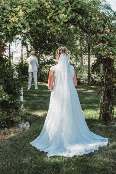 First Look in Full Bloom Bride in a white gown approaches a groom in a garden setting.
